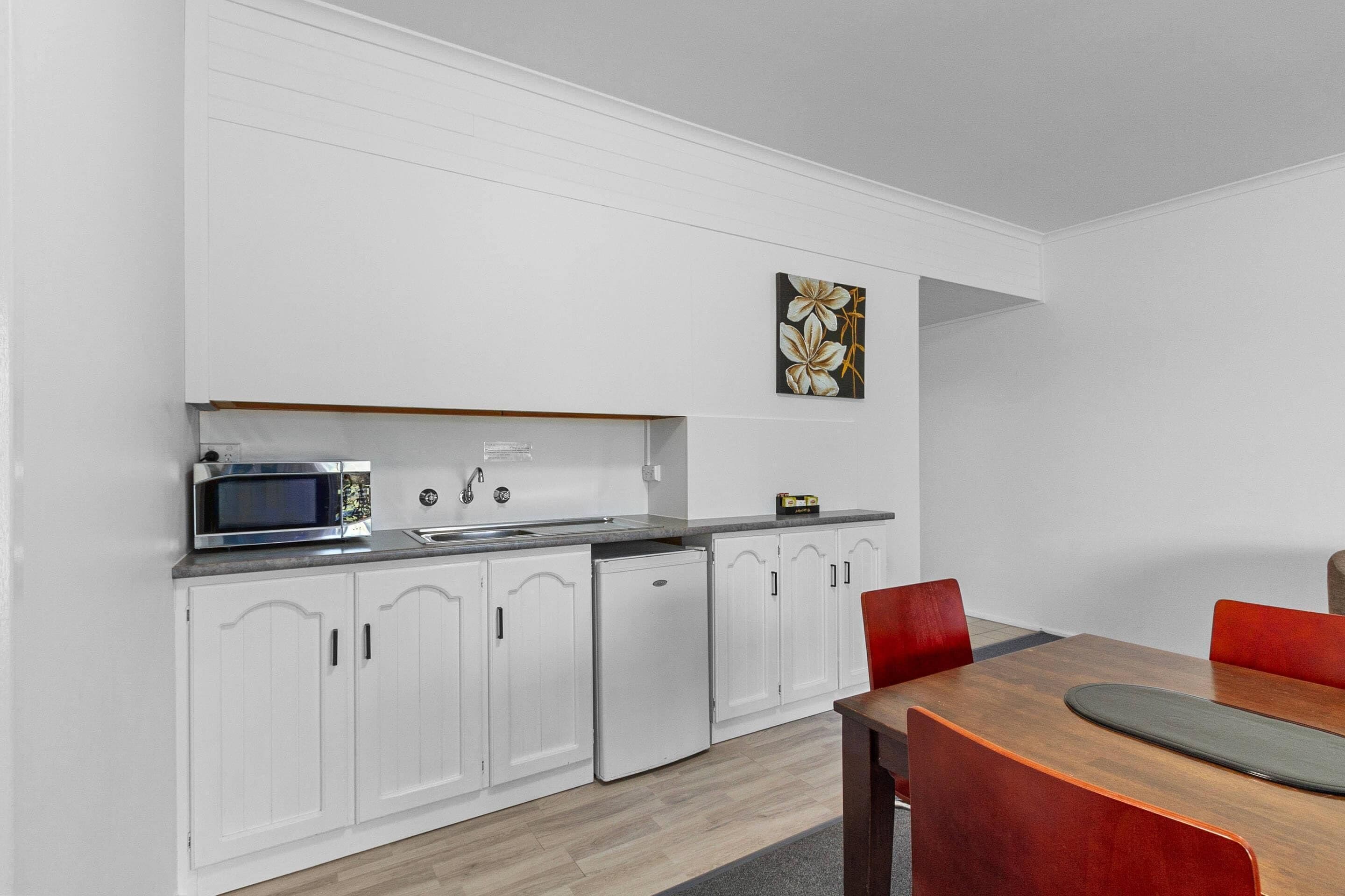 Una cocina moderna con armarios blancos, un microondas, un fregadero y una zona de comedor con una mesa de madera y sillas rojas.