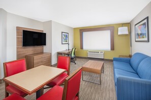 Une chambre d’hôtel compacte comprenant un coin repas, un canapé, un téléviseur fixé au mur et une fenêtre avec des stores.