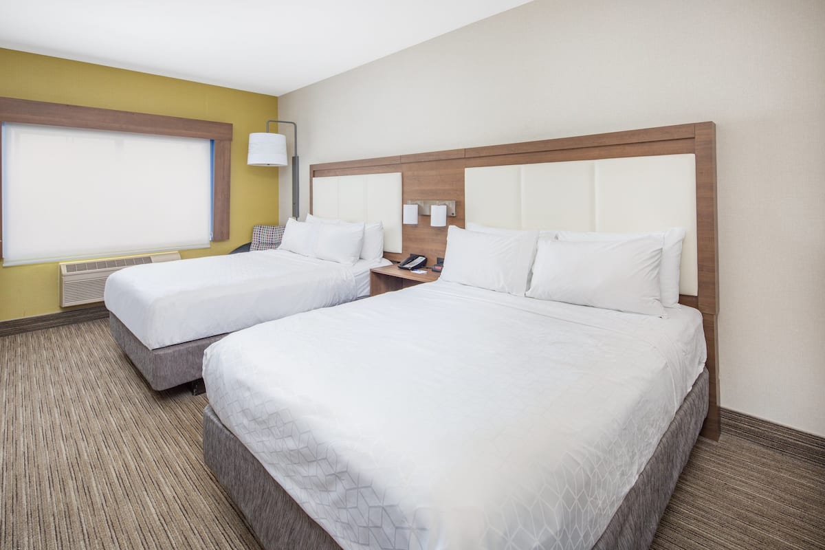 A hotel room with two beds, a window with blinds, a bedside table with a lamp, and a wall-mounted phone.