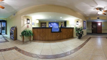 Zona de recepción con mostrador curvo, un letrero azul y una recepcionista. Hay plantas en macetas y un piso de baldosas con un borde decorativo.