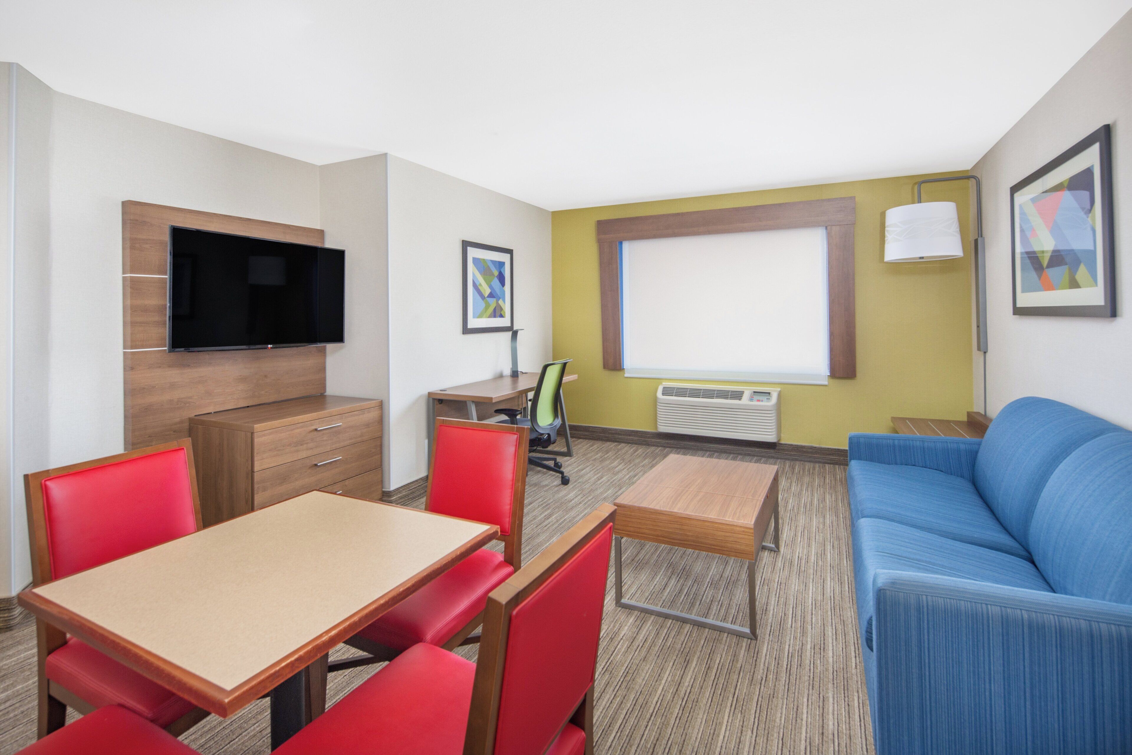 A hotel room with a TV mounted on the wall, a desk with a chair, a sofa, and a window with blinds.
