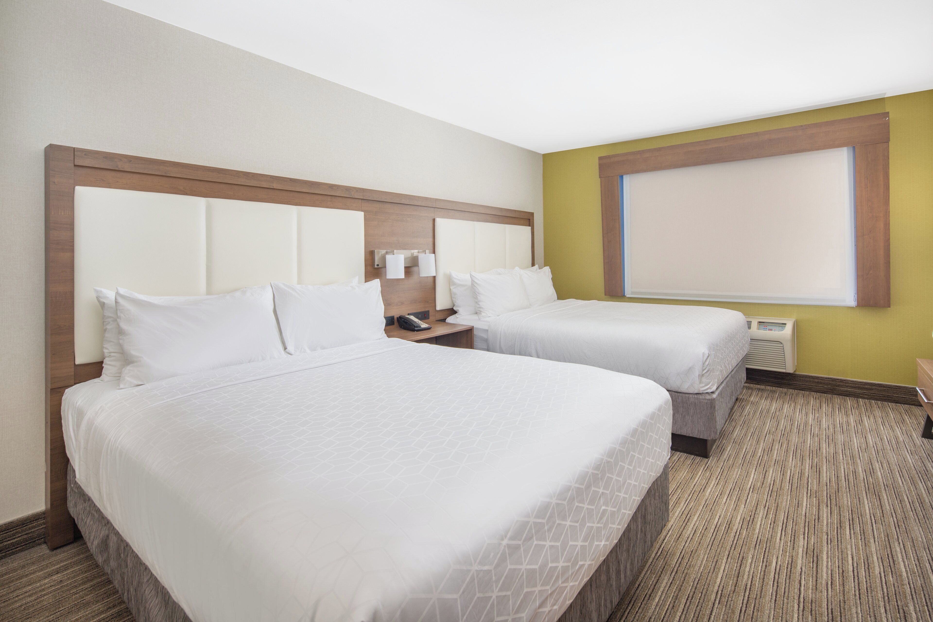 Quarto de hotel com duas camas, cabeceira de madeira, mesa de cabeceira com telefone e janela com persianas.
