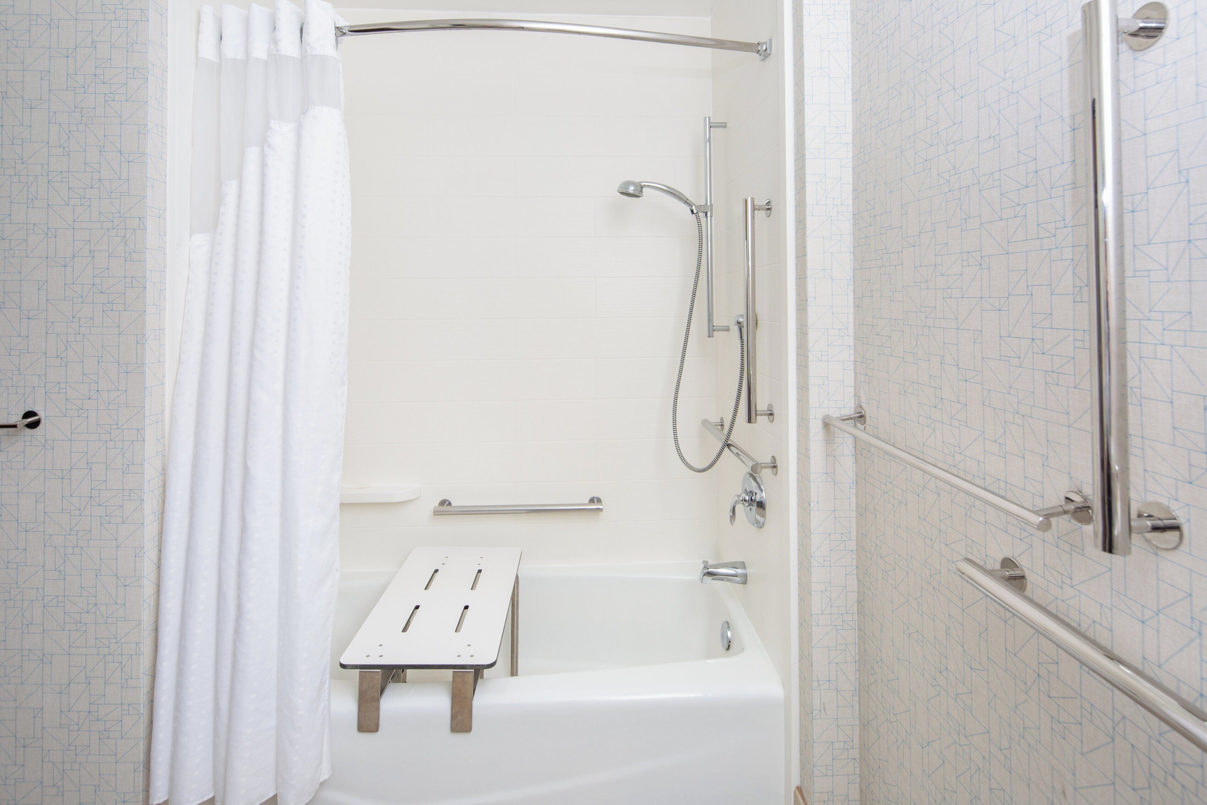 Eine barrierefreie Dusche mit Duschabtrennung aus Glas, einer Handbrause und einem zusätzlichen Duscharmaturen, sowie einer kleinen Stufe zur Erleichterung des Ein- und Ausstiegs.