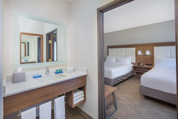 Une chambre d’hôtel avec un grand miroir, un lavabo, une coiffeuse, deux lits et une table de chevet avec des lampes.