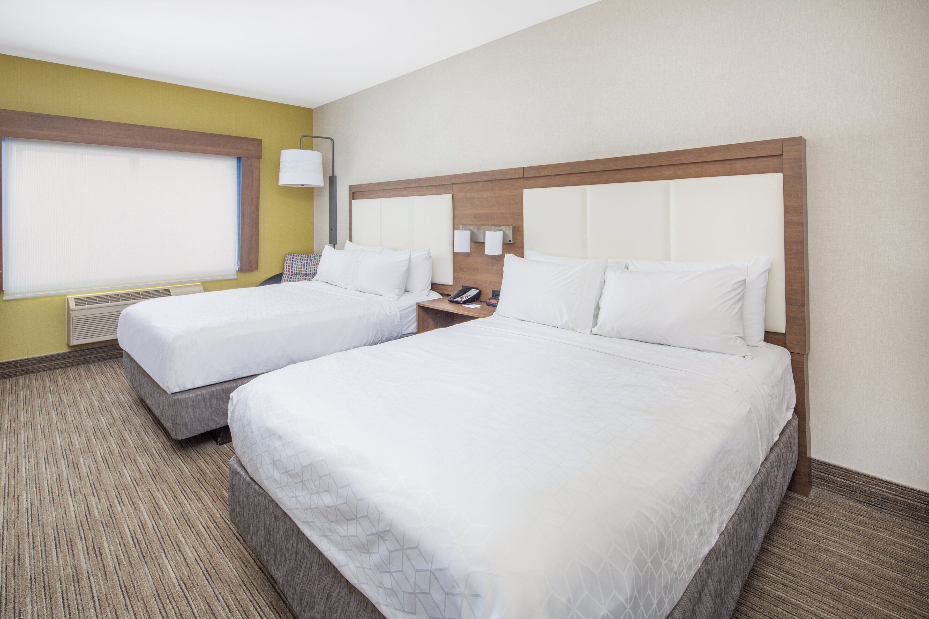 A hotel room with two beds, a window with blinds, a bedside table with a lamp, and a wall-mounted phone.