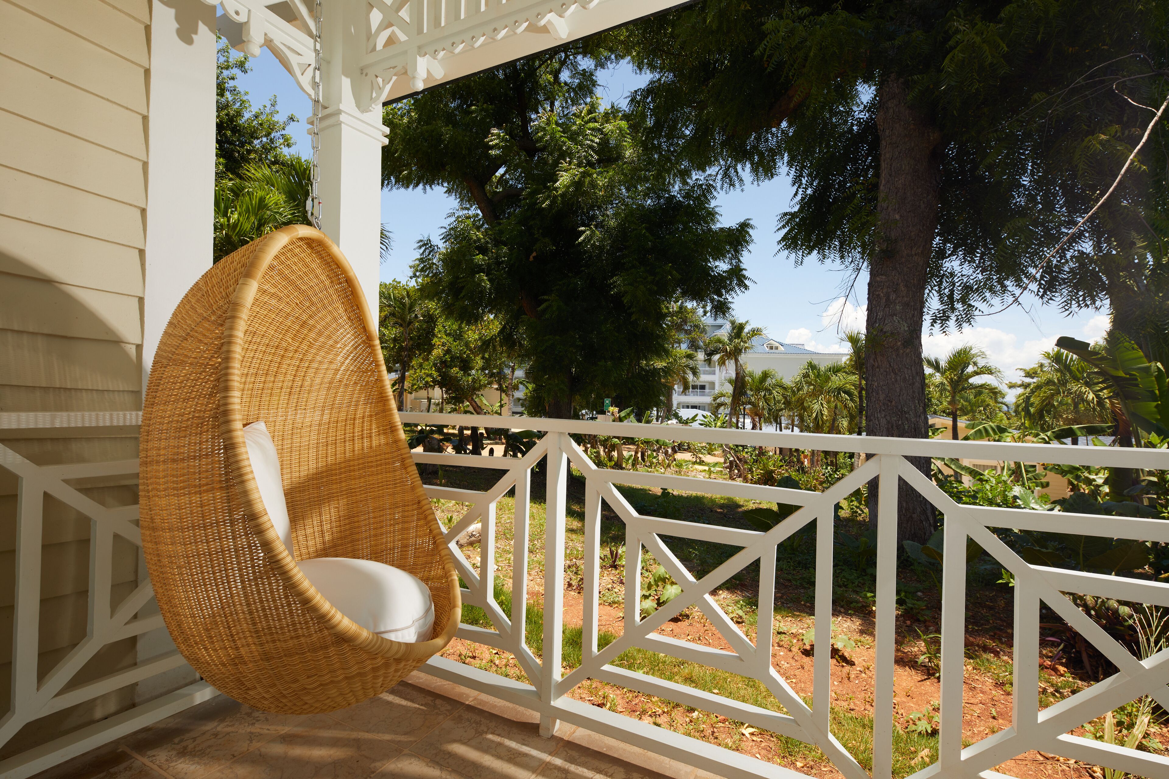A wicker chair on a porch with a white railing, overlooking a garden with trees and a distant view of a building.