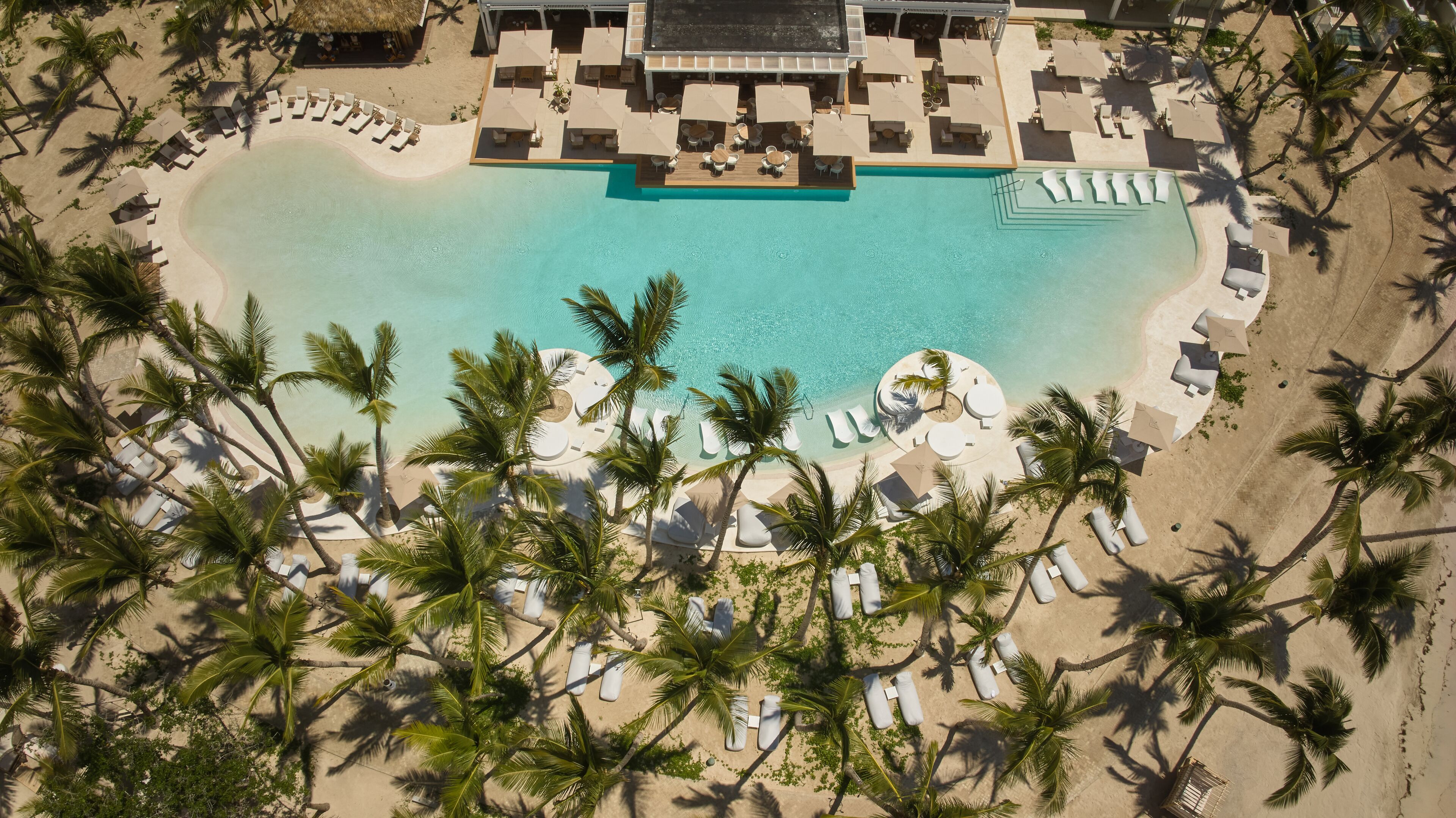 An aerial view of a resort with a central swimming pool, surrounded by palm trees and sun loungers.