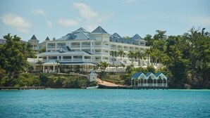 Un ampio hotel a più piani con facciata bianca e tetto blu, affacciato su un mare cristallino con palme e un pontile.