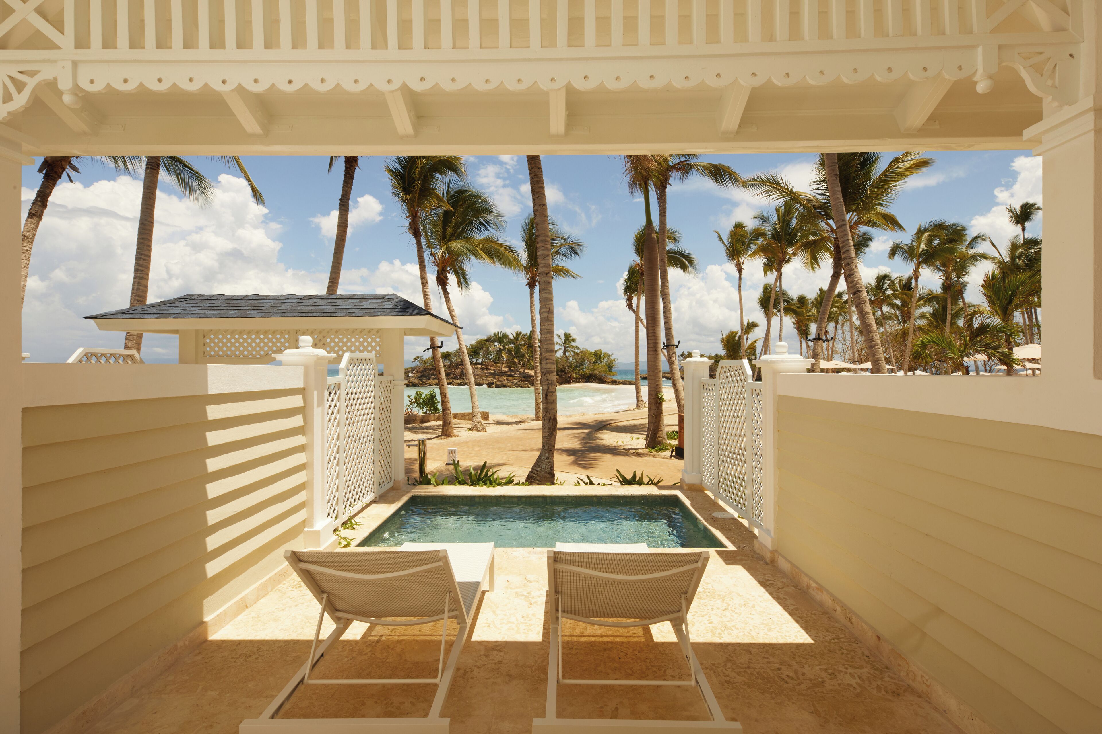 A pool area with two lounge chairs, a view of a beach with palm trees, and a clear sky.