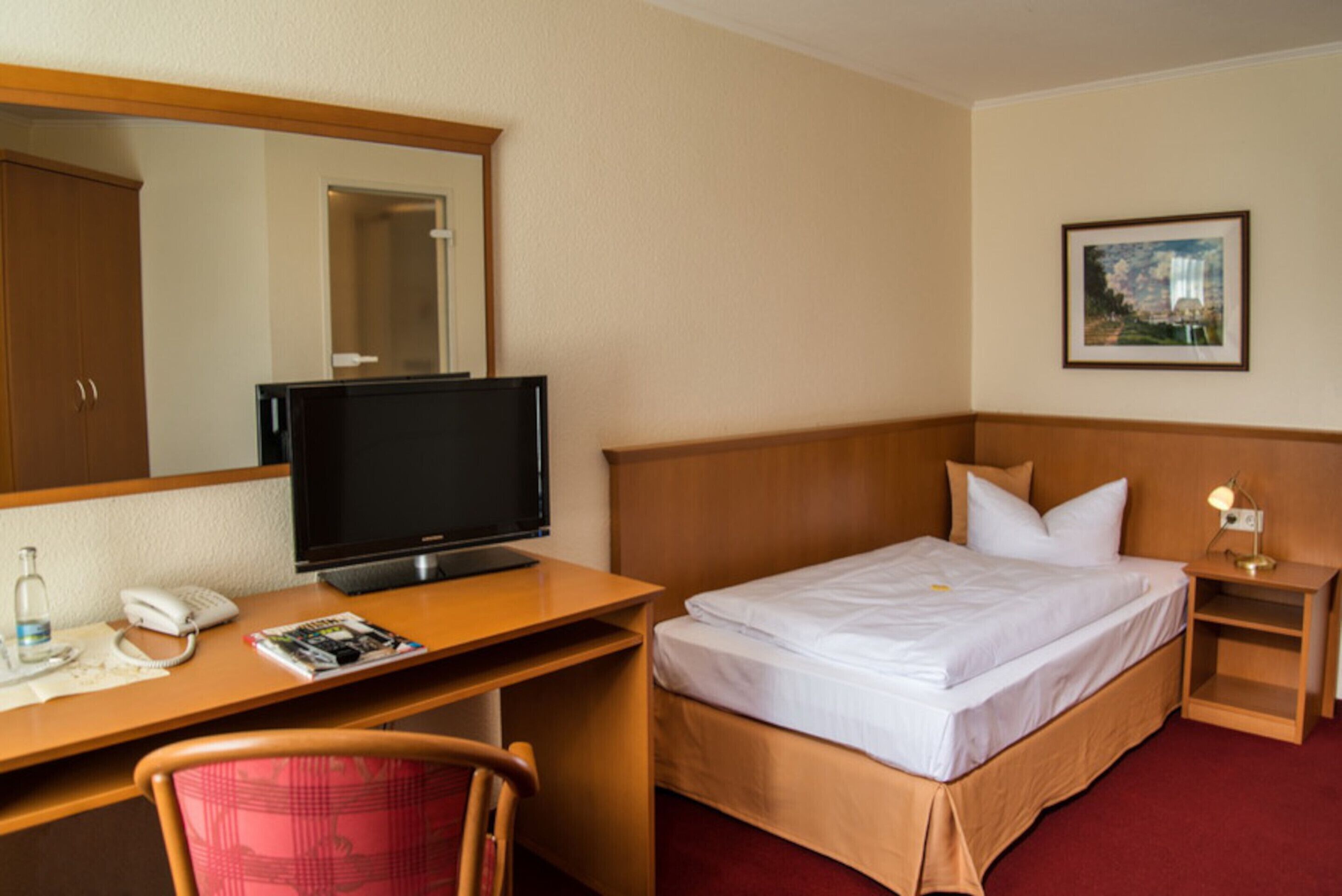 A hotel room with a bed, a desk with a computer, a chair, a bedside table with a lamp, and a framed picture on the wall.