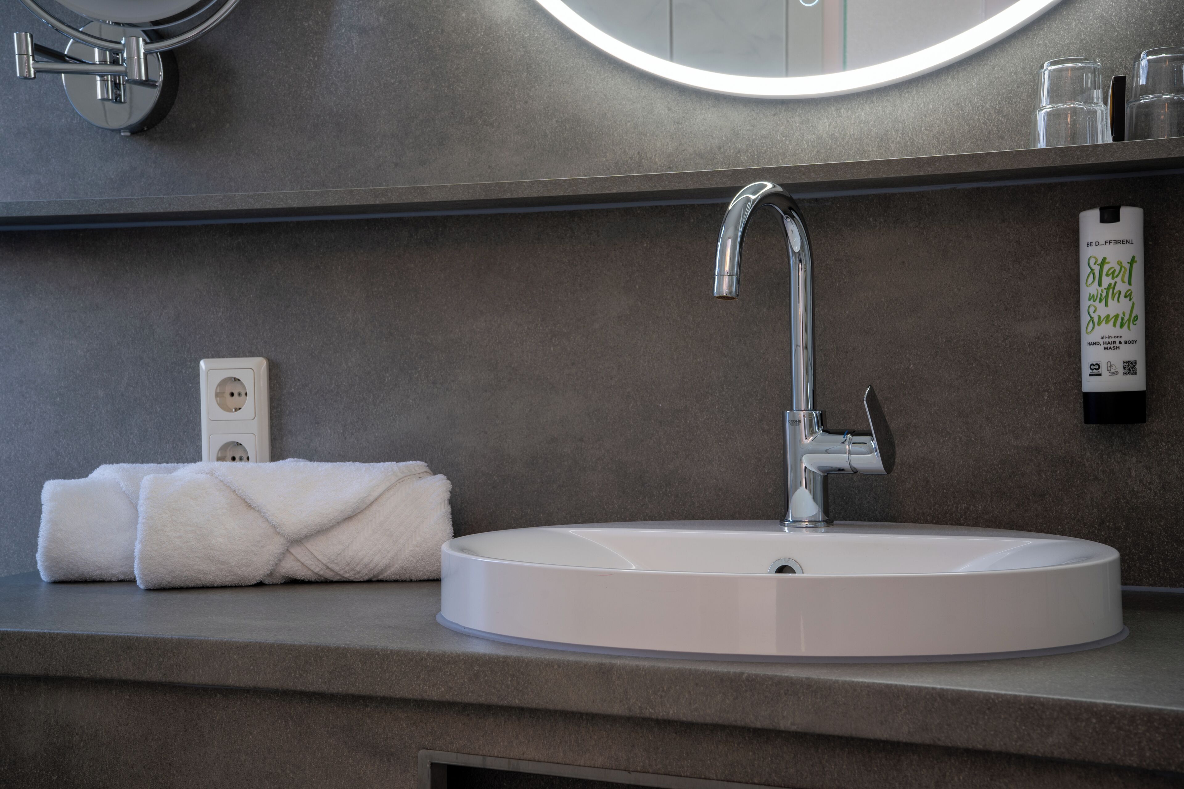 Un baño moderno con un lavabo blanco redondeado, un grifo cromado, una toalla, un espejo y un dispensador de jabón en la pared.
