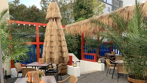 An outdoor seating area with wicker furniture, a thatched umbrella, and a red and blue fence.