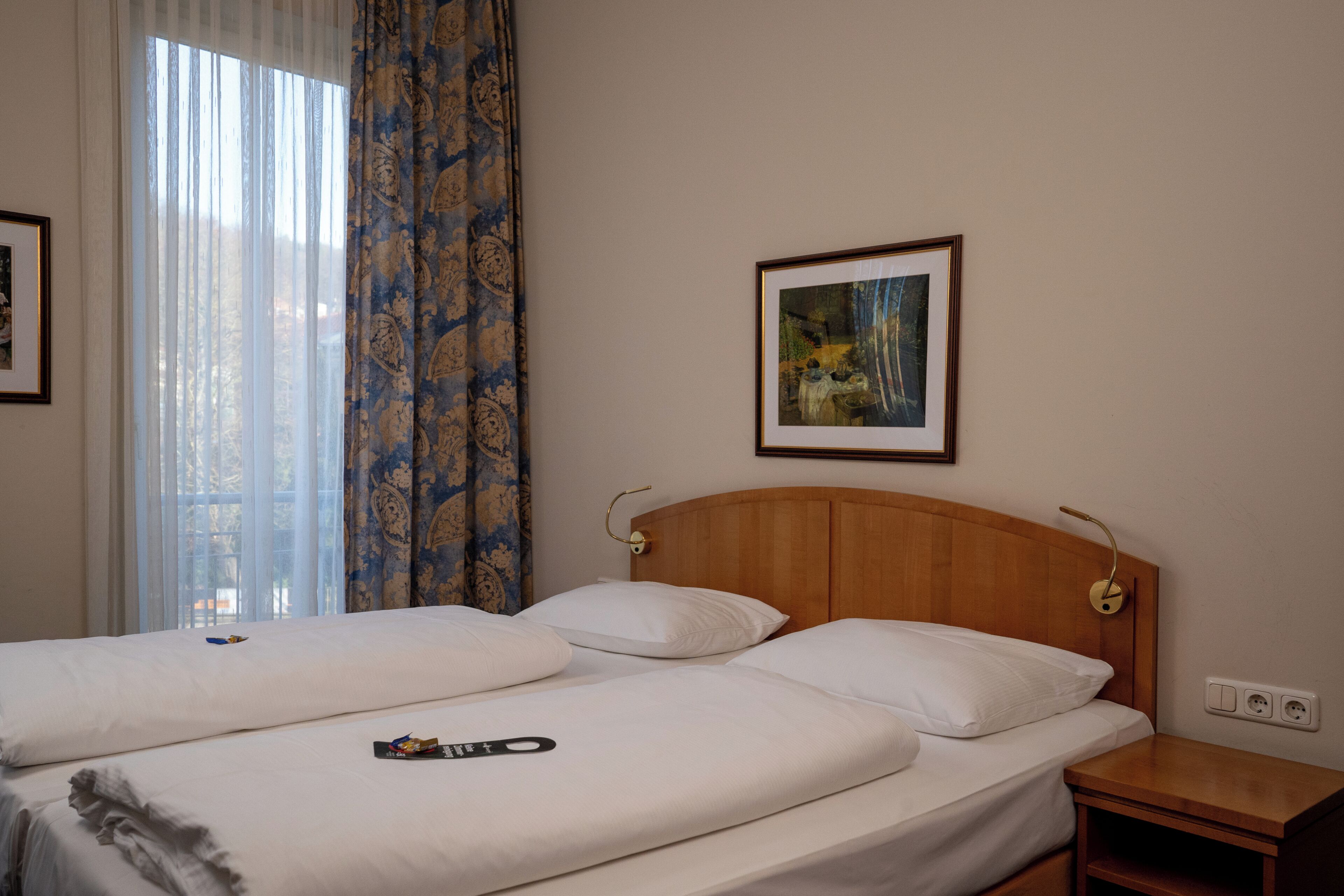 Ein Hotelzimmer mit zwei Betten, einem hölzernen Kopfteil, einem Nachttisch, einem gerahmten Bild an der Wand und einem Fenster mit durchsichtigen Vorhängen.