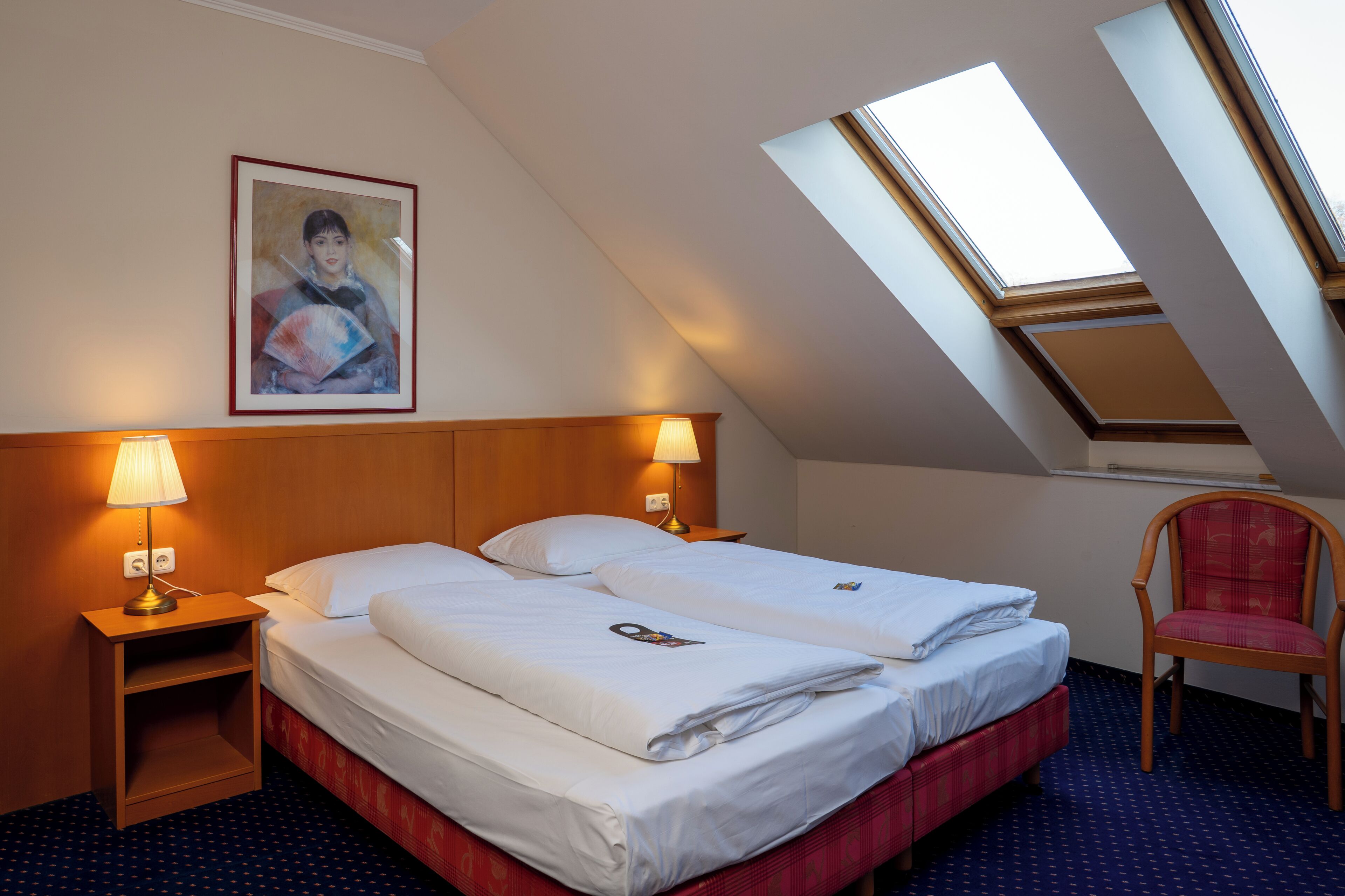 A hotel room with two beds, a nightstand with a lamp, a chair, a painting on the wall, and skylights.