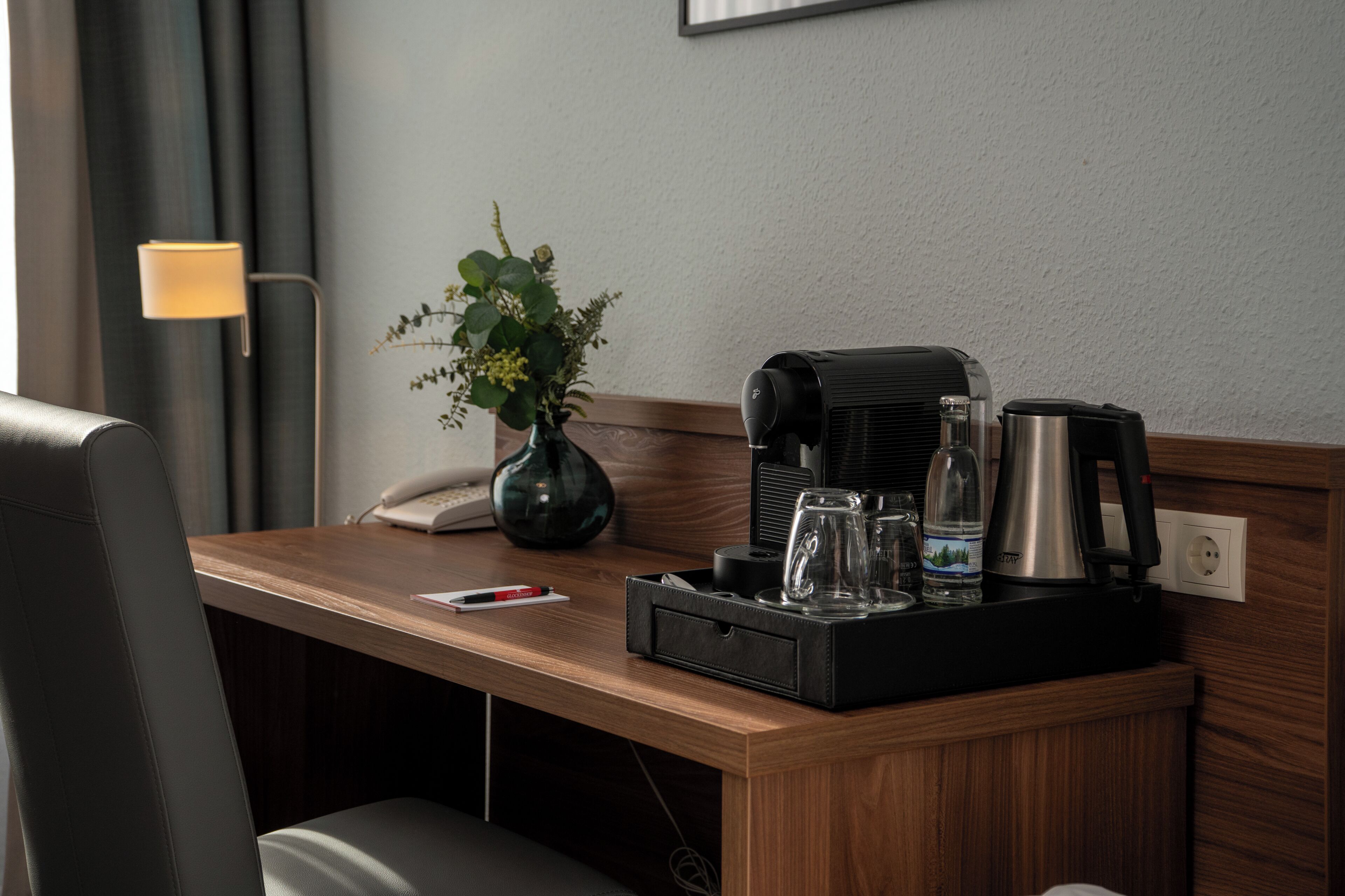 Escritorio de hotel con cafetera, botella de agua, vaso y un florero con follaje.