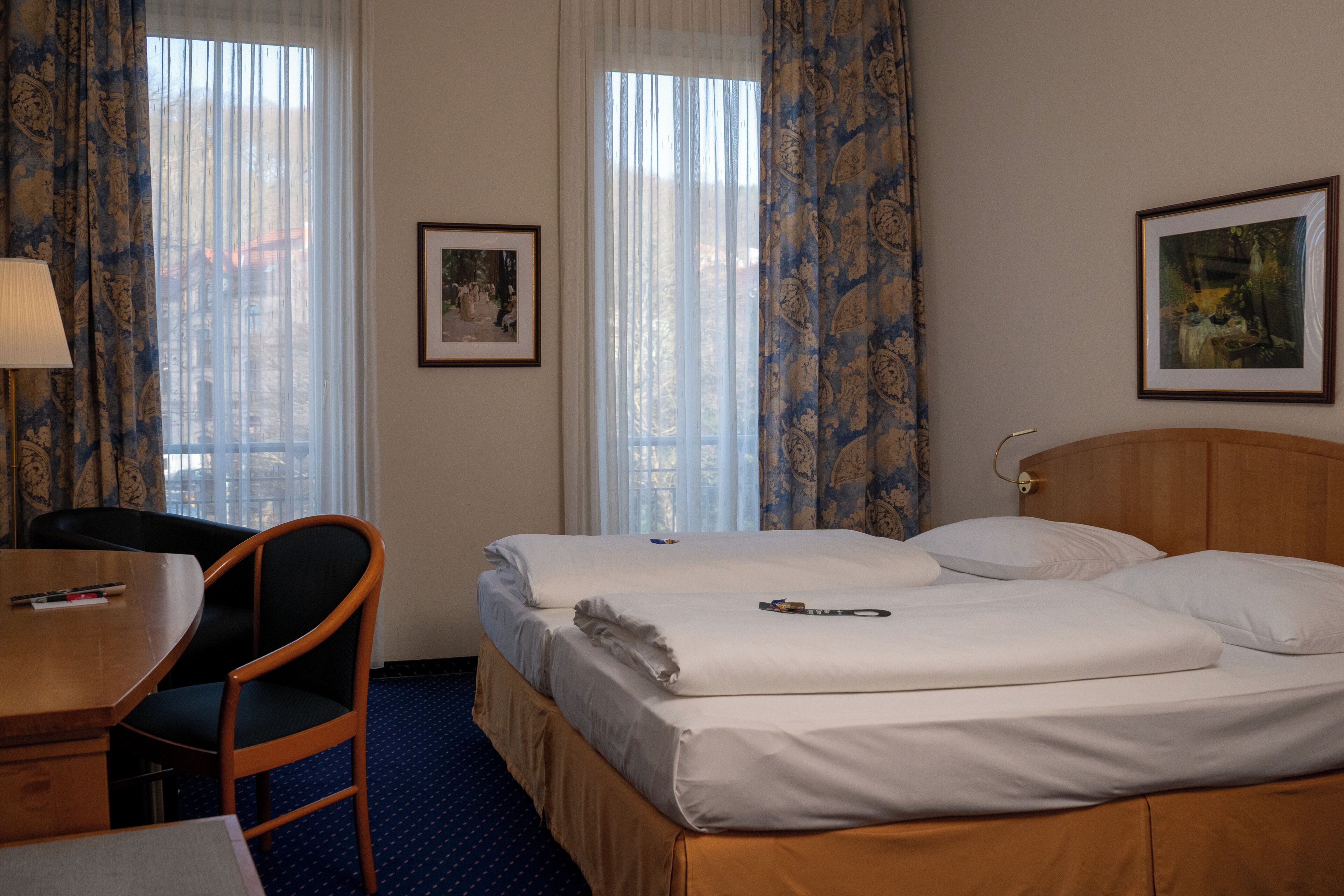A hotel room with a bed, a desk, a chair, and two windows with curtains.