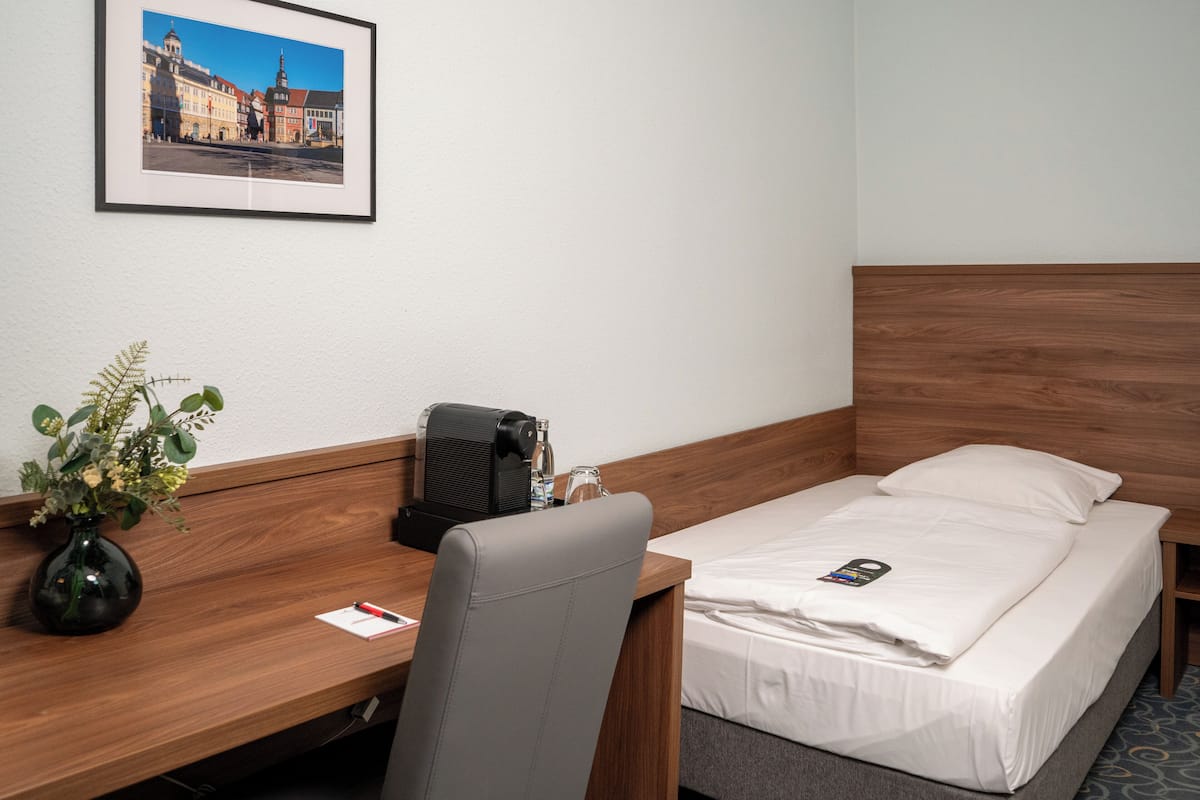 Habitación de hotel con una cama, un escritorio, una silla, una cafetera, un florero con flores y un cuadro en la pared.