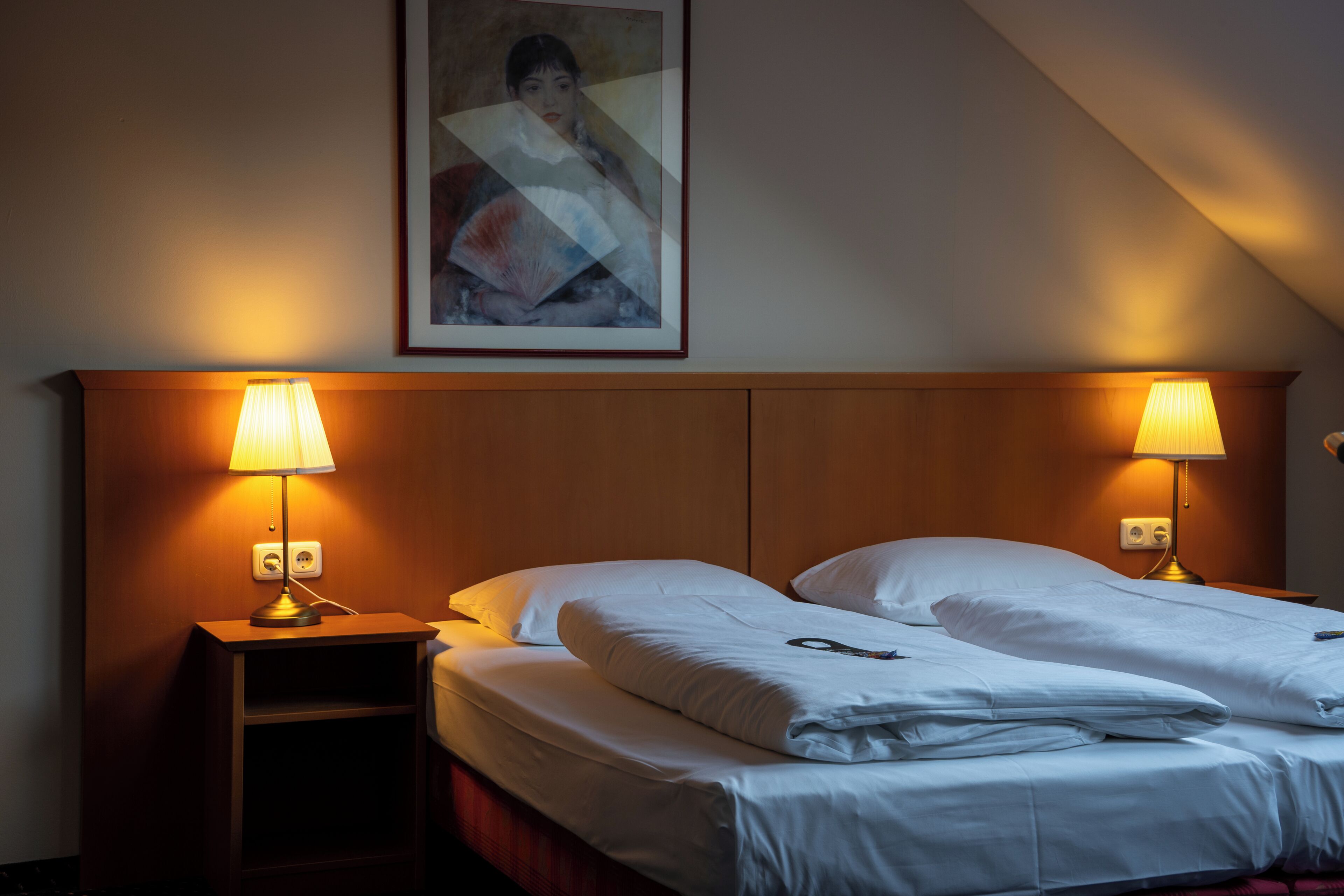 Habitació d'hotel amb un capçal de llit de fusta, dues làmpades de nit, un llit amb roba blanca i un quadre a la paret.