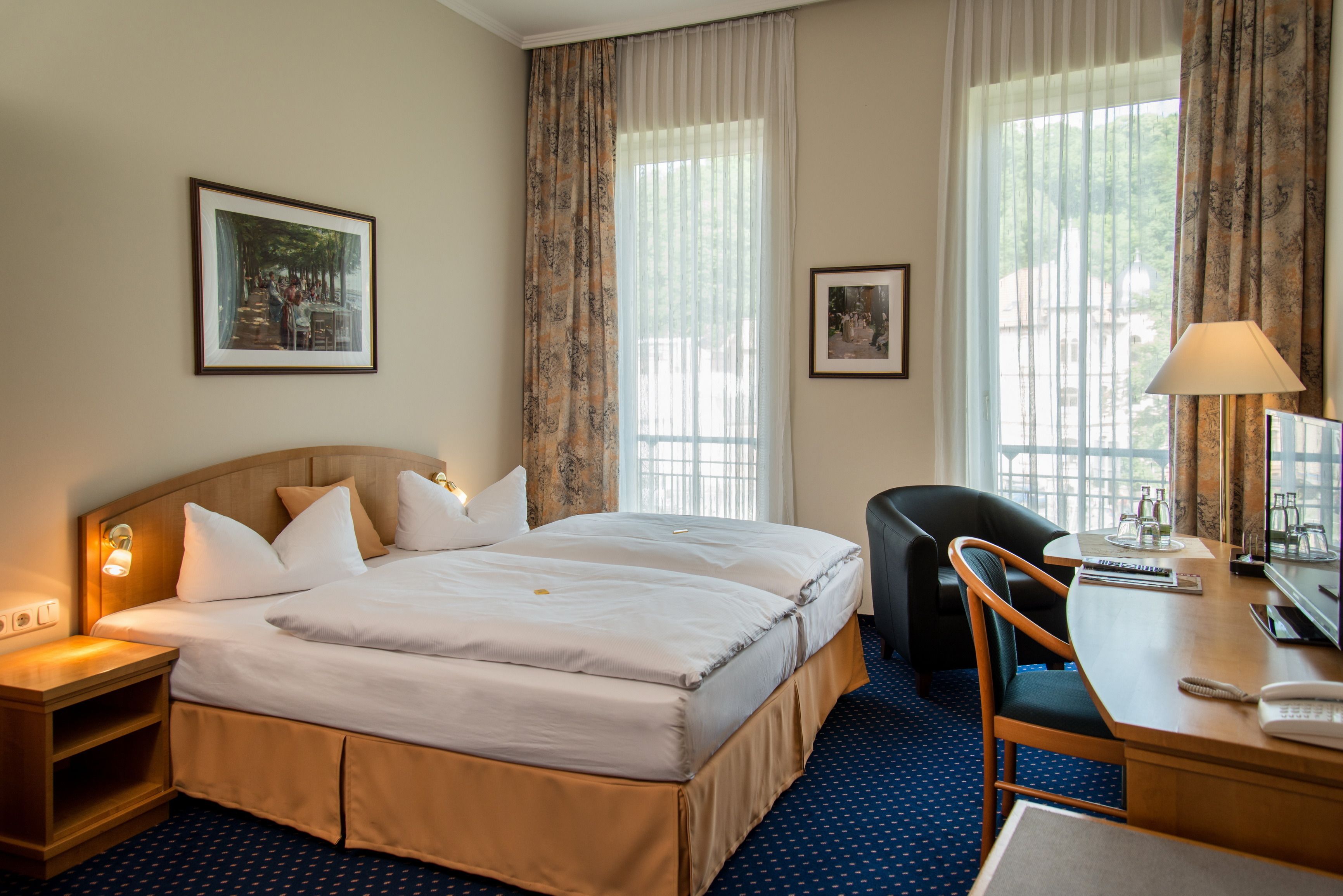Una habitación de hotel con una cama doble, un escritorio con lámpara, una silla y dos cuadros en la pared.