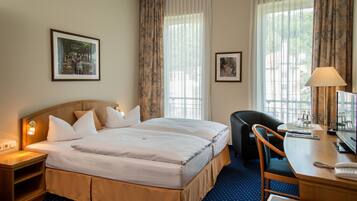 A hotel room with a double bed, a desk with a lamp, a chair, and two framed pictures on the wall.