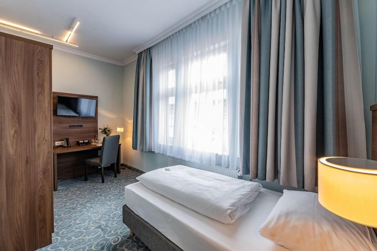 A hotel room with a bed, a desk with a chair, a television, and a window with curtains.
