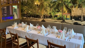 A restaurant with a long table set for dining, tropical-themed murals on the walls, and a view of a beach.