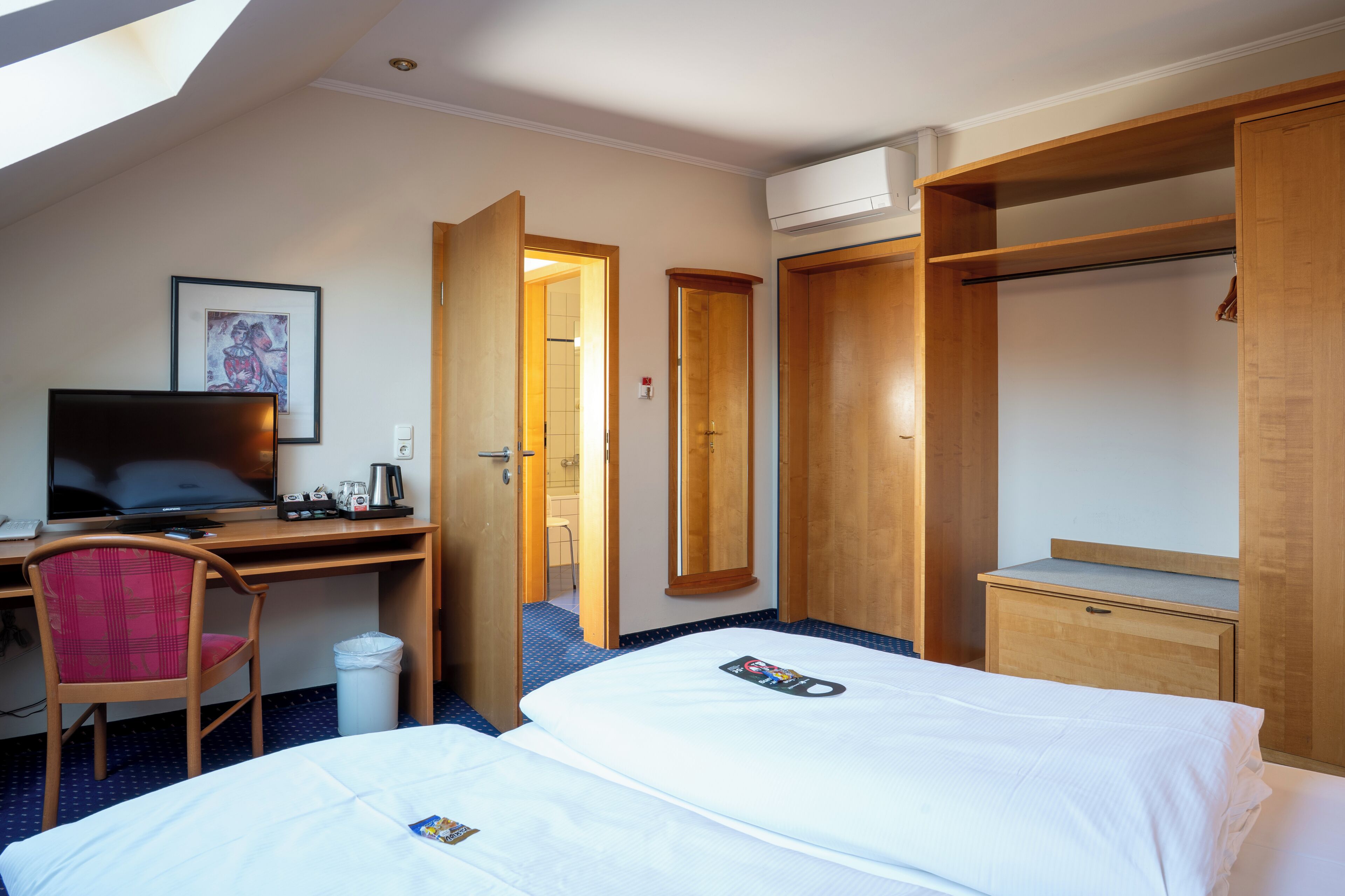 A hotel room with two beds, a desk with a computer, a chair, a wardrobe, and an air conditioning unit.
