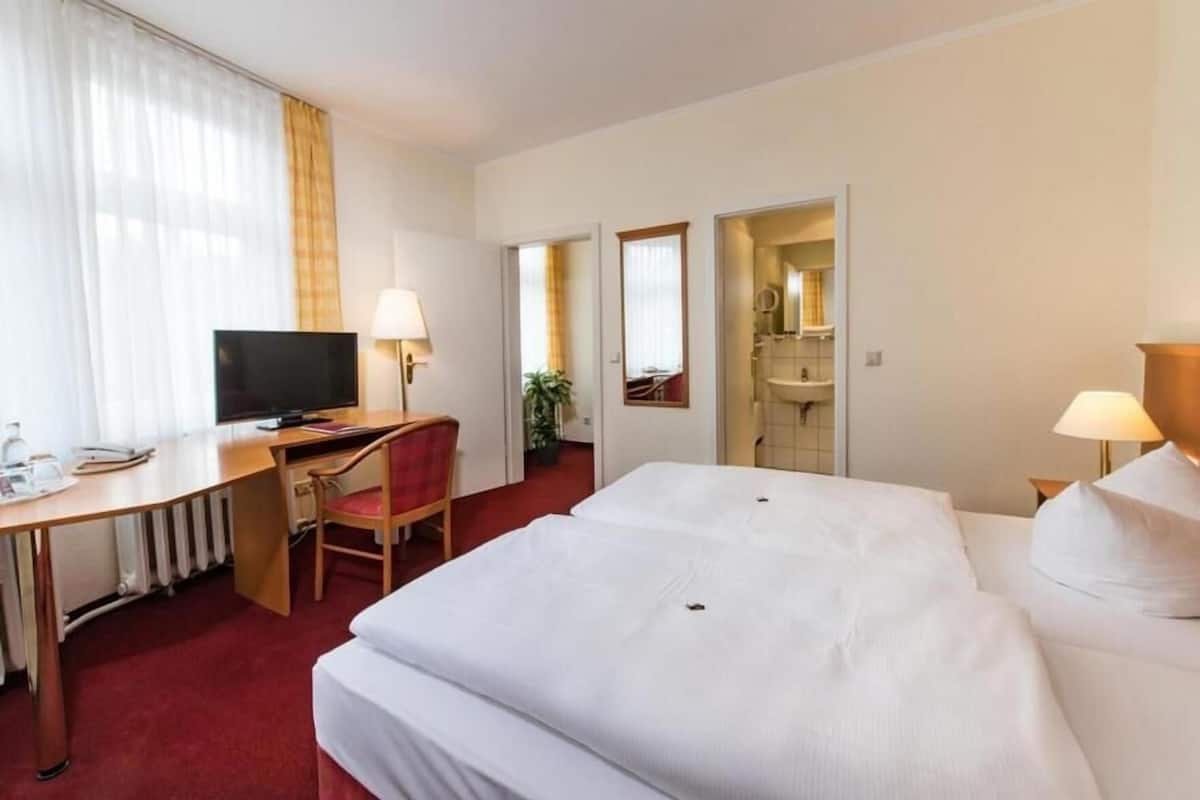 Une chambre d’hôtel avec un lit, un bureau doté d’une télévision, une chaise, une lampe et une salle de bain visible à travers une porte ouverte.
