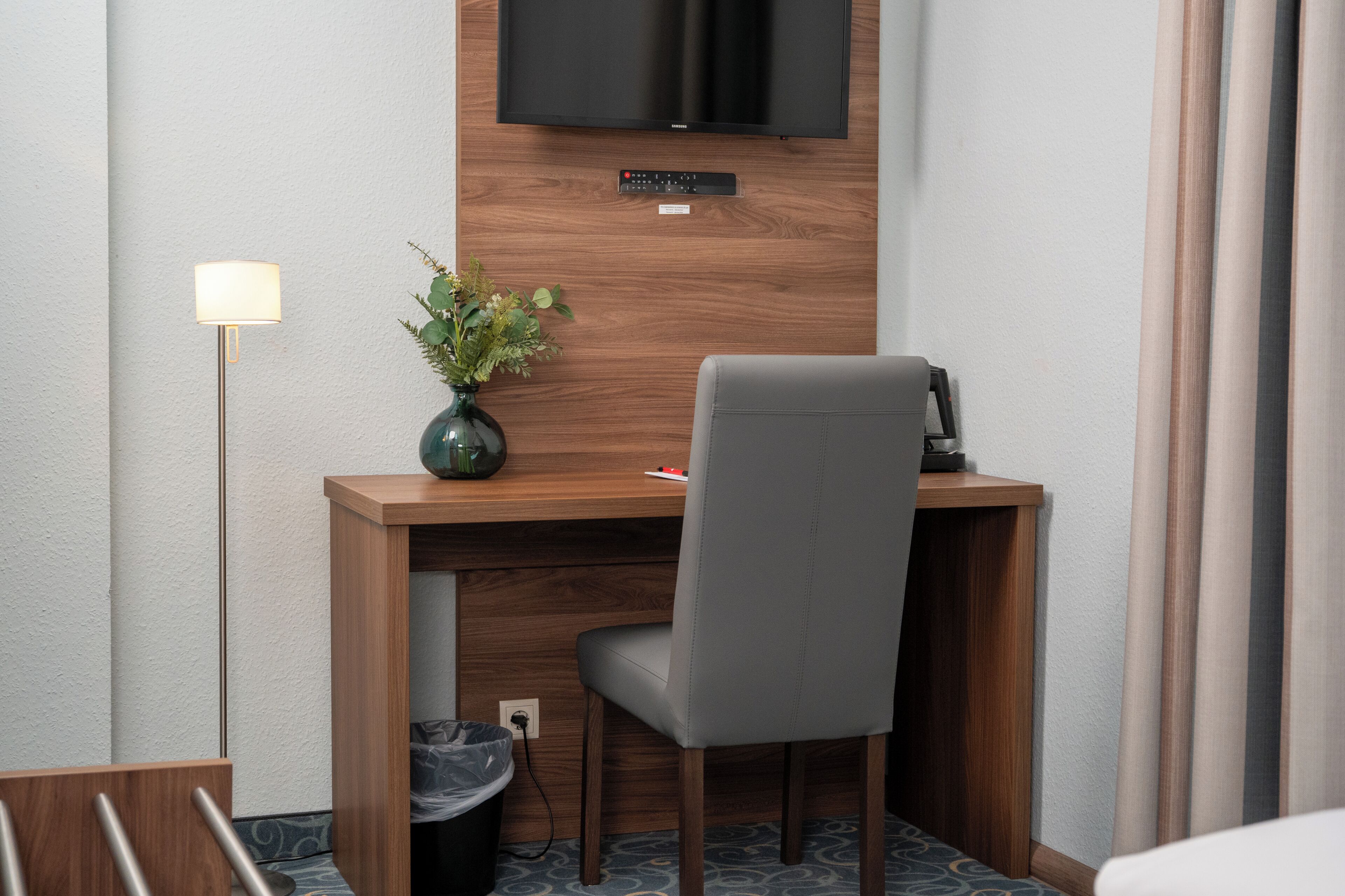 A hotel room with a wooden desk, a grey chair, a television mounted on the wall, a small plant on the desk, and a floor lamp.