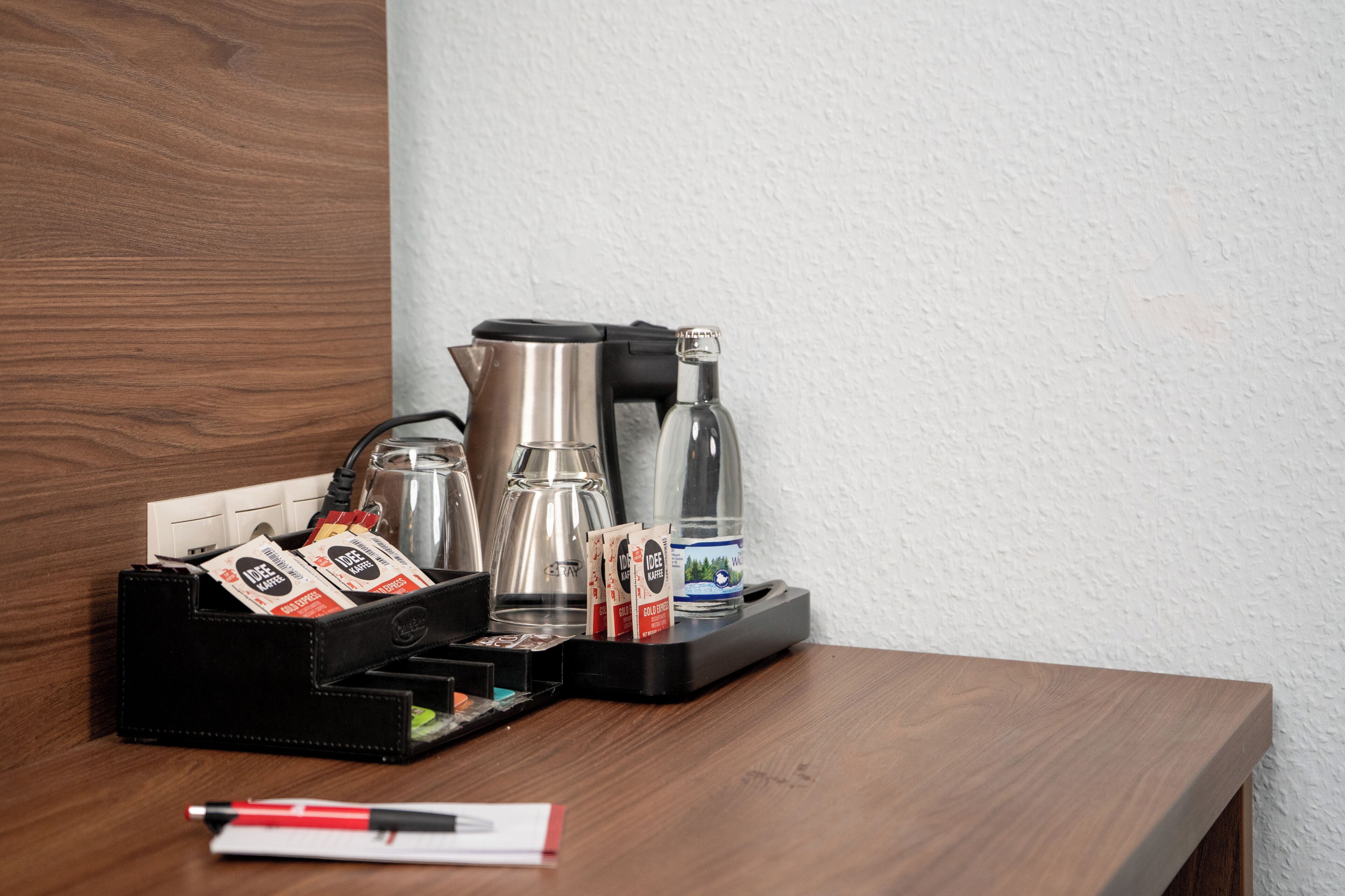 Un coin café comprenant une machine à café, une verseuse en verre, un verre d’eau et des dosettes de café, le tout posé sur une table en bois devant un mur blanc.