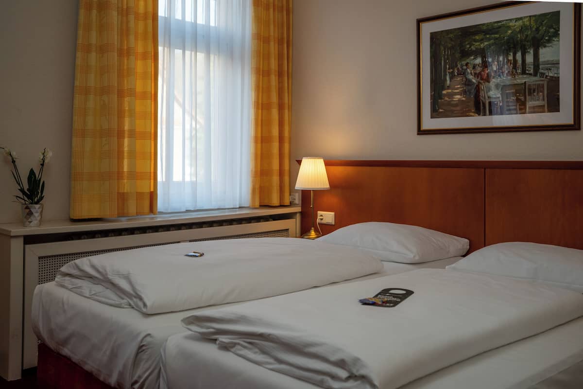 Una habitación de hotel con dos camas, una ventana con cortinas amarillas, un cuadro en la pared y una lámpara de noche.