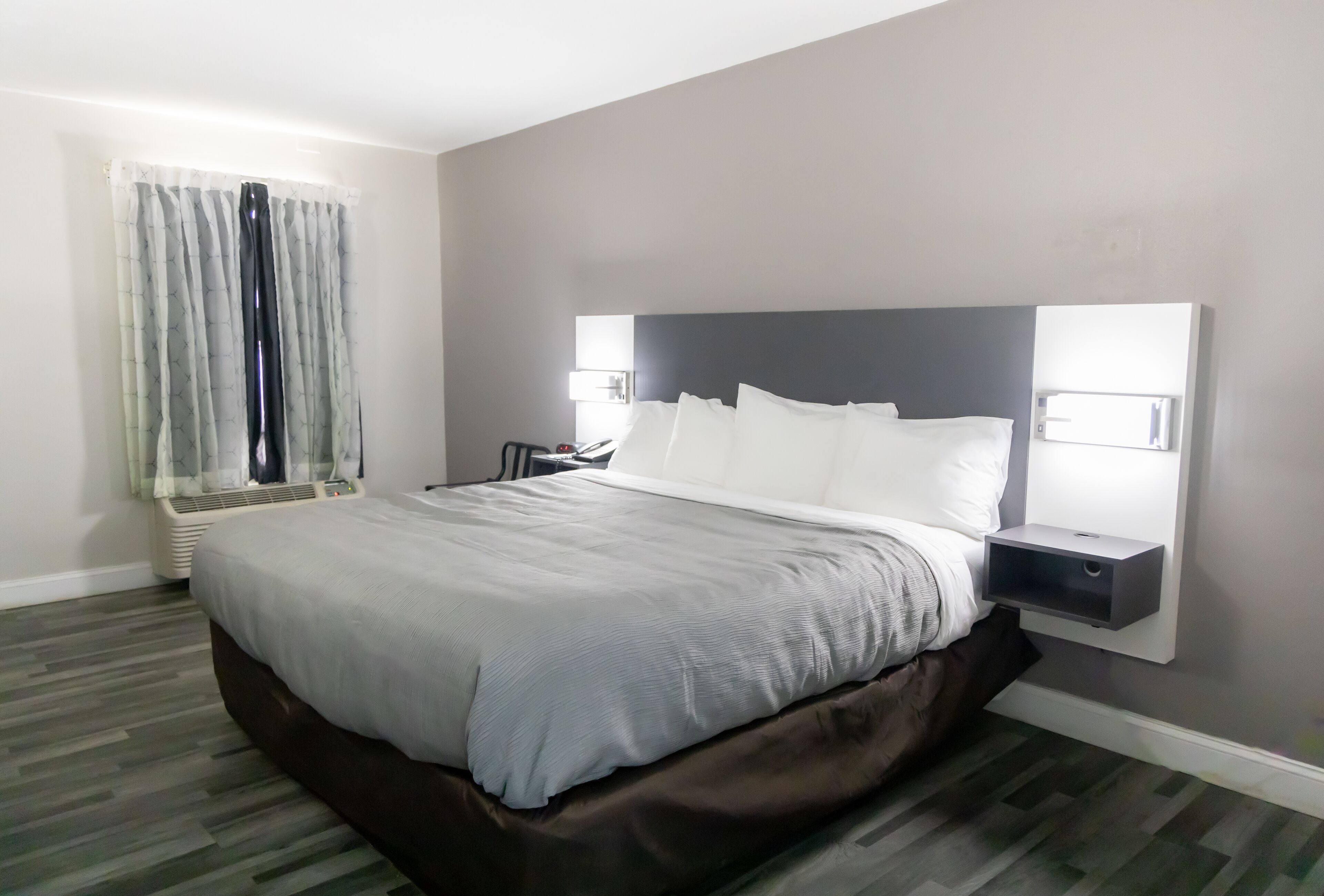 Una habitación de hotel moderna con una cama grande, paredes grises y una mesita de noche empotrada con una lámpara.