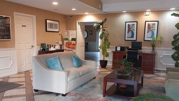 A spacious living area with a white sofa, a coffee table, and a ceiling fan. There are potted plants and framed artwork on the walls. - Quality Inn Carrollton Medical Center (Carrollton)