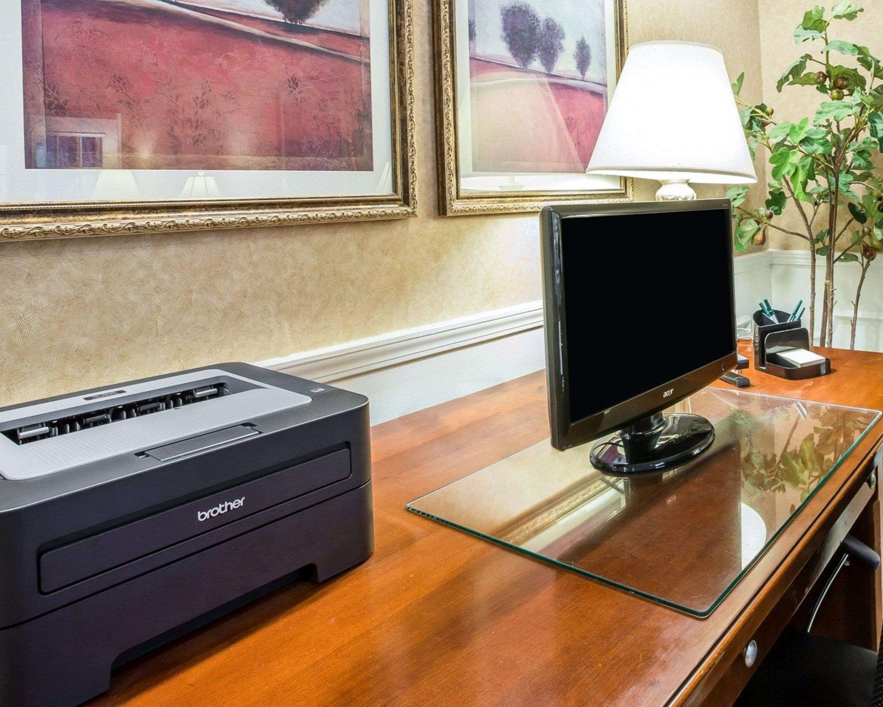 A printer and monitor on a wooden desk with a glass top, framed artwork on the wall, and a lamp.