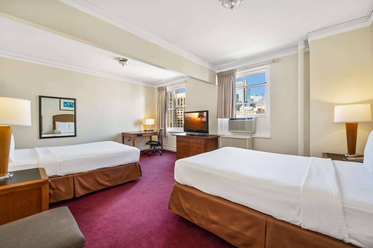 A hotel room with two beds, a desk, a television, and a window with a city view.