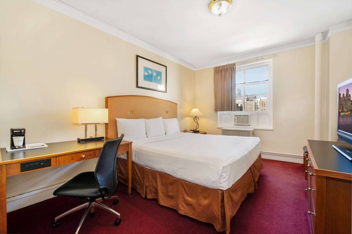 A hotel room with a bed, desk, chair, and a window with a city view.