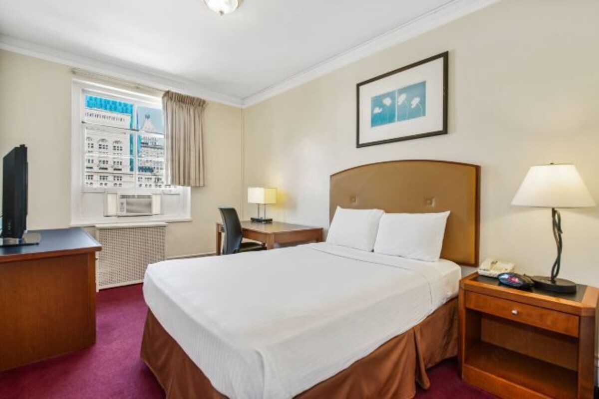 Habitación de hotel con cama, escritorio, silla, mesita de noche, lámpara, cuadro enmarcado y ventana con vista a edificios.