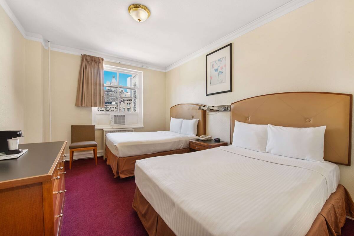 A hotel room with two beds, a desk, a chair, a window with a view of buildings, and a framed picture on the wall.