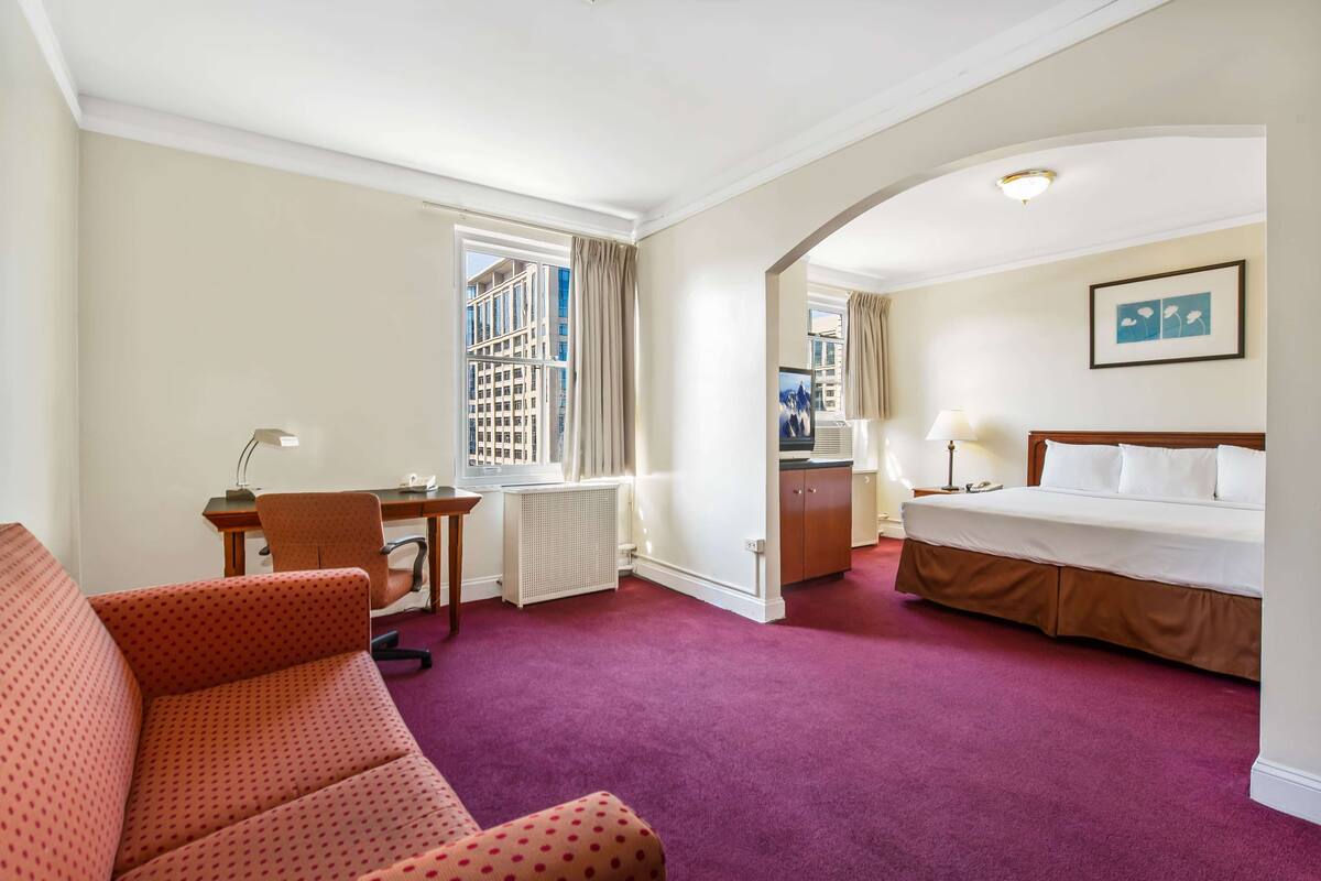 A hotel room with a bed, a desk with a chair, a sofa, and a view of a building through the window.