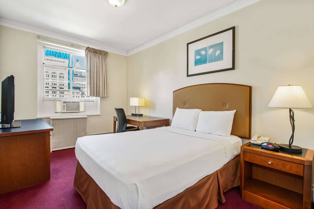 A hotel room with a large bed, a desk with a computer, a chair, a bedside table with a lamp, and a window with a cityscape view.