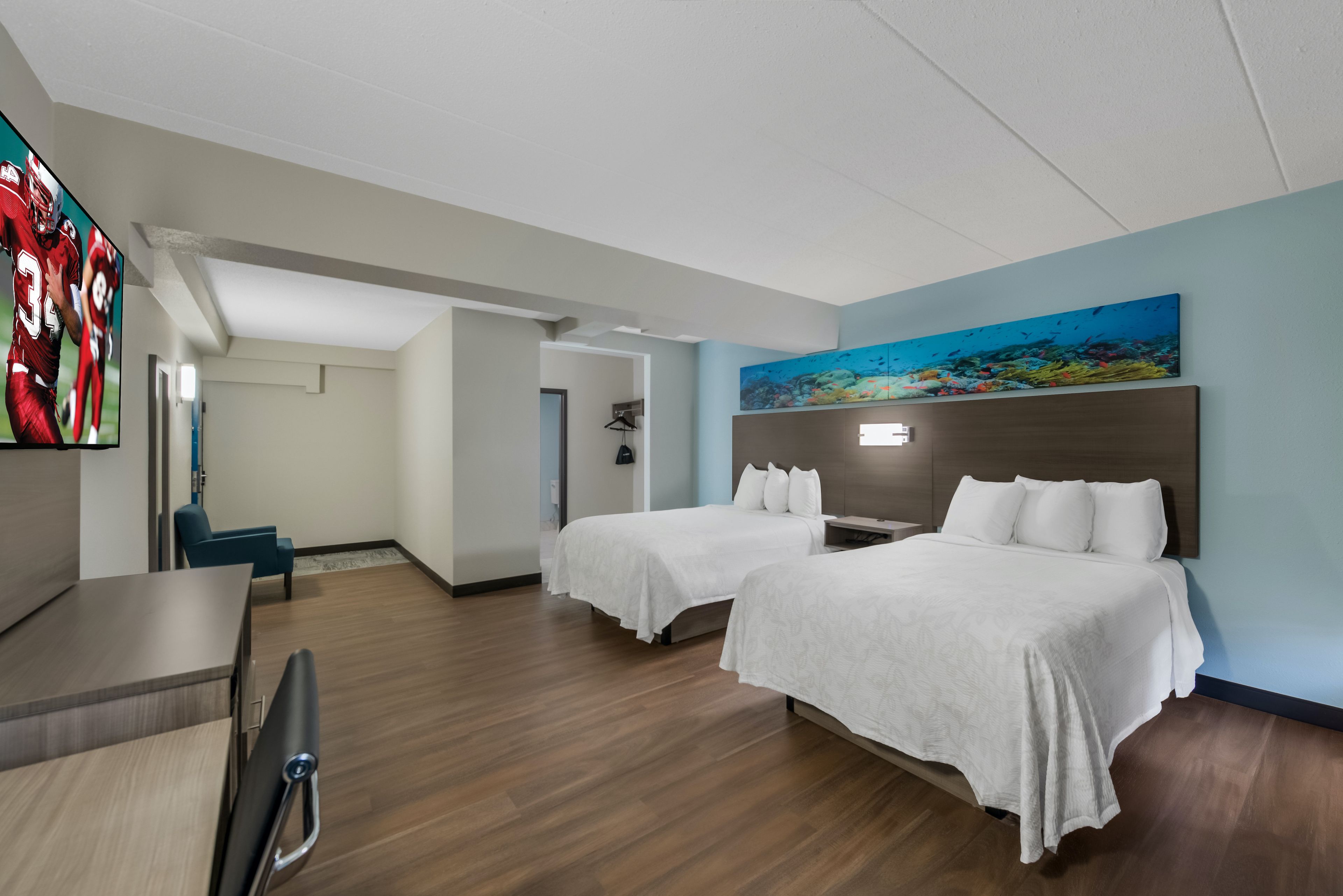 A hotel room with two beds, a desk, a chair, and a TV displaying sports.
