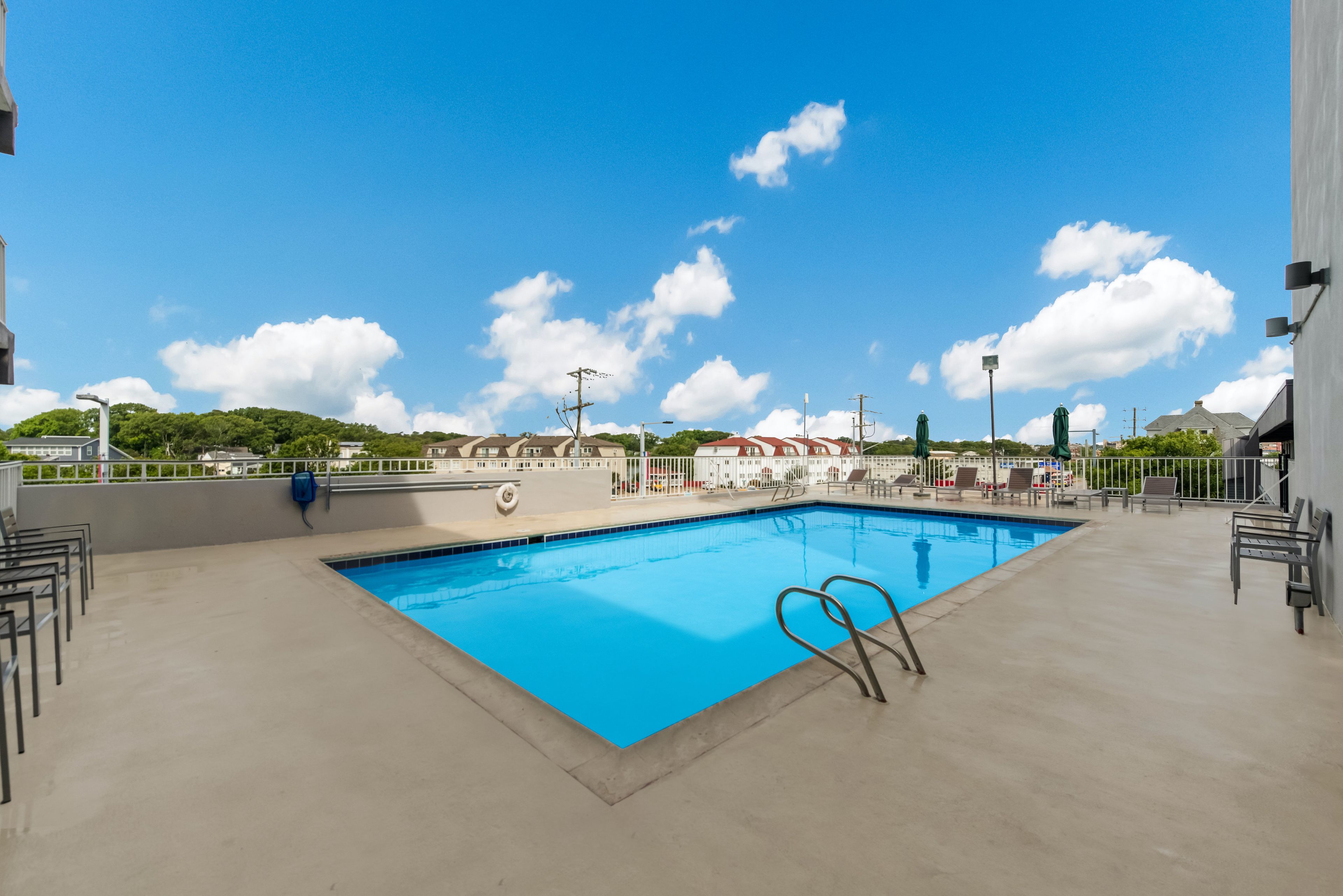 Piscina retangular com superfície de água azul cristalina, rodeada por um deck de concreto com grades de metal e algumas cadeiras de descanso.