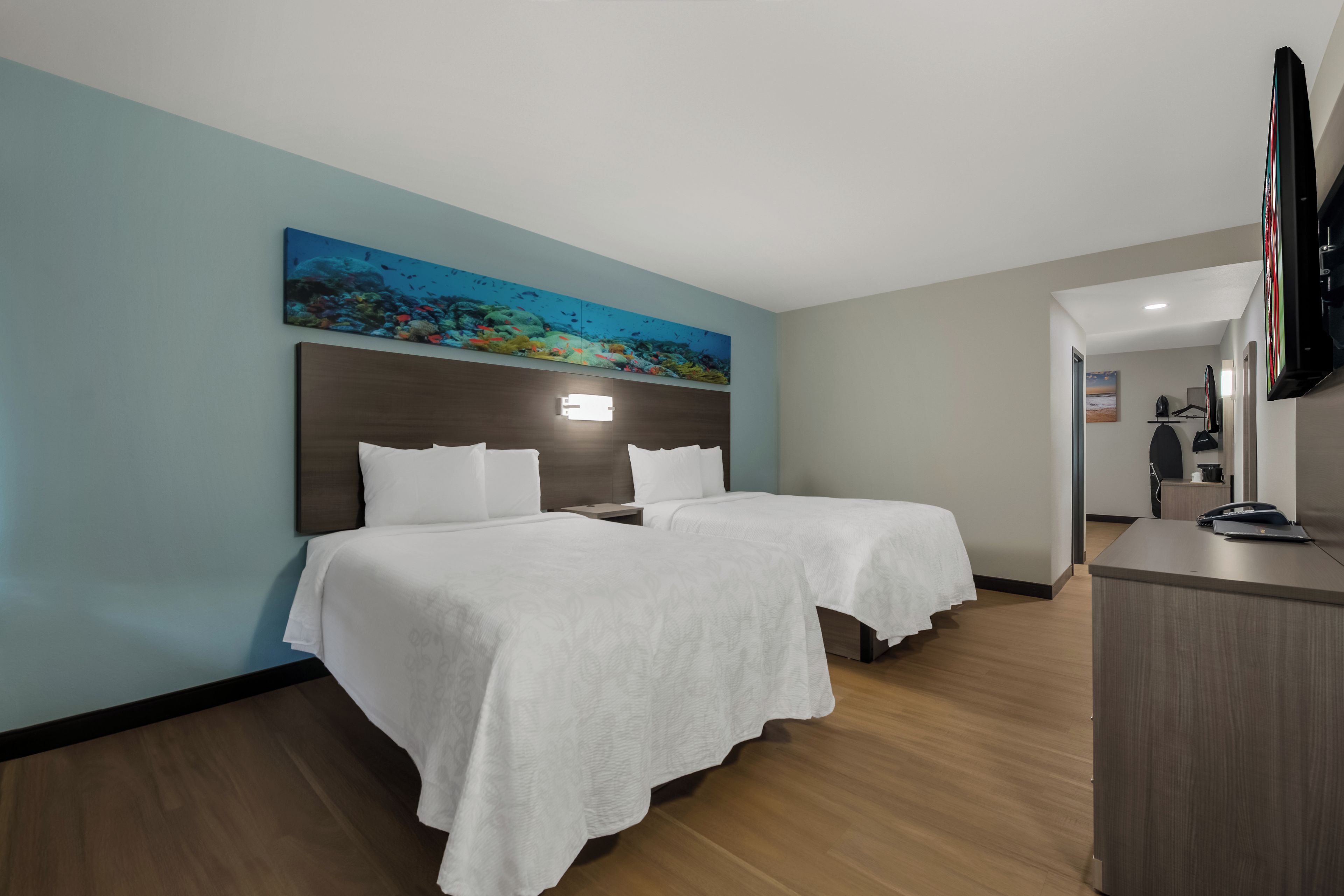 A hotel room with two beds, a large painting of an underwater scene, a flat-screen TV, and a wooden desk with a telephone.