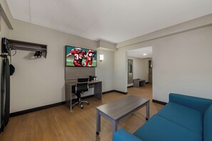A modern living room with a flat-screen TV, a desk, a blue sofa, and a coffee table. - Red Roof Inn PLUS+ & Suites Virginia Beach – Seaside (Virginia Beach)