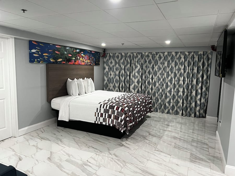 Een slaapkamer met een bed, hoofdeinde, een kunstwerk aan de muur, gordijnen met een patroon en een betegelde vloer.