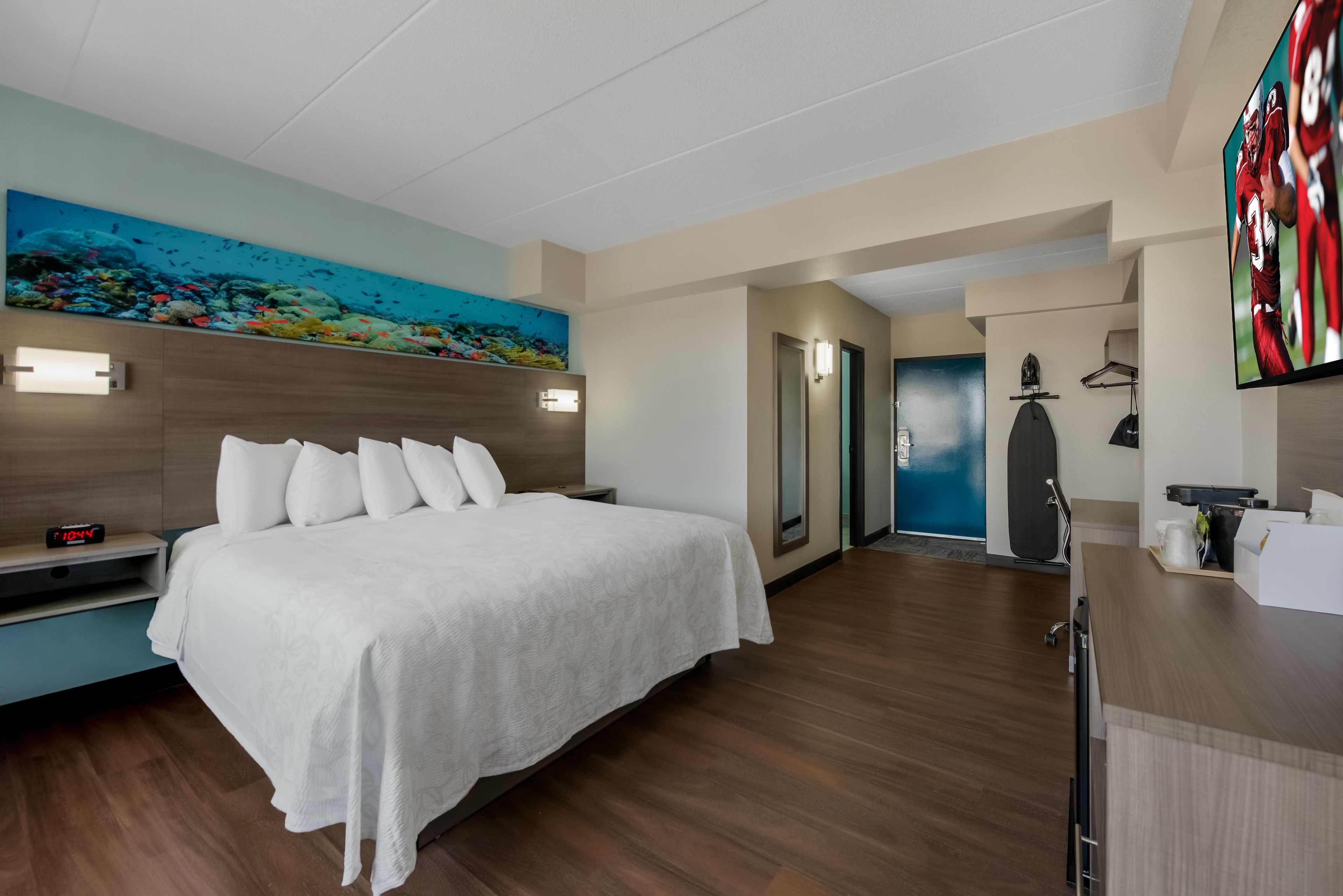 A hotel room with a bed, a painting of an underwater scene, a desk with a computer, and a door leading to another room.