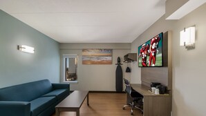 A modern living room with a blue sofa, a wooden coffee table, a desk with a chair, a flat-screen TV, and a painting of a sunset. - Red Roof Inn PLUS+ & Suites Virginia Beach – Seaside (Virginia Beach)