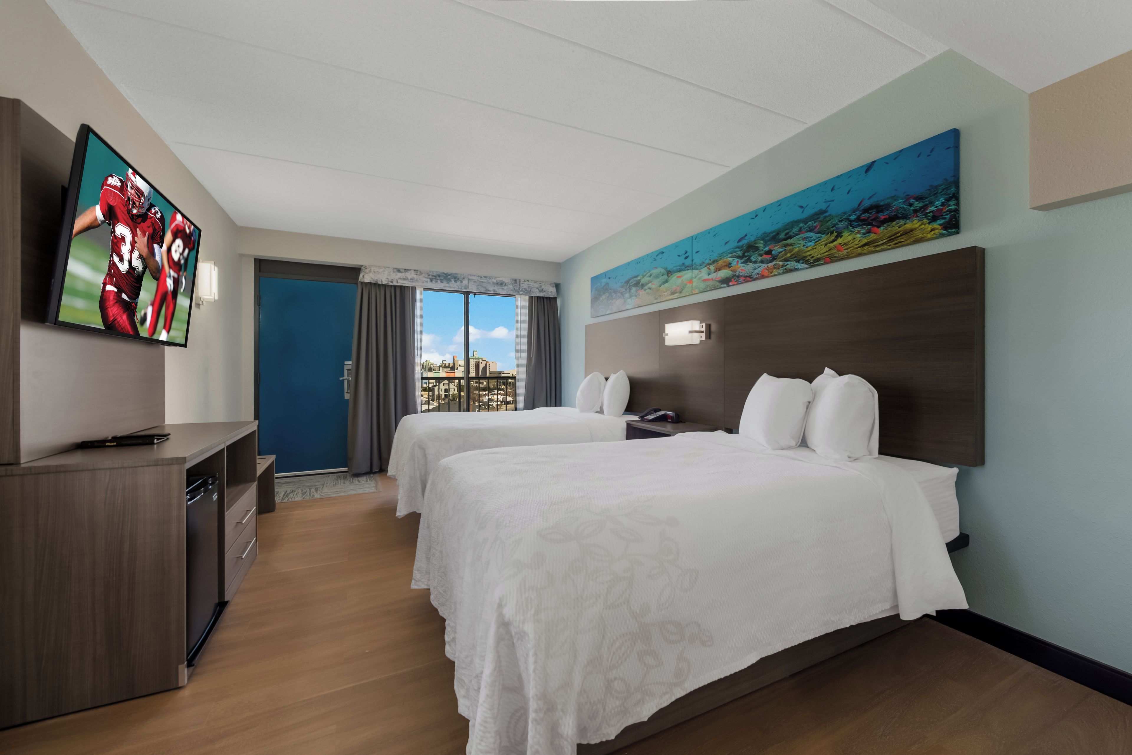 A hotel room with two beds, a TV, a painting, and a view of the city.