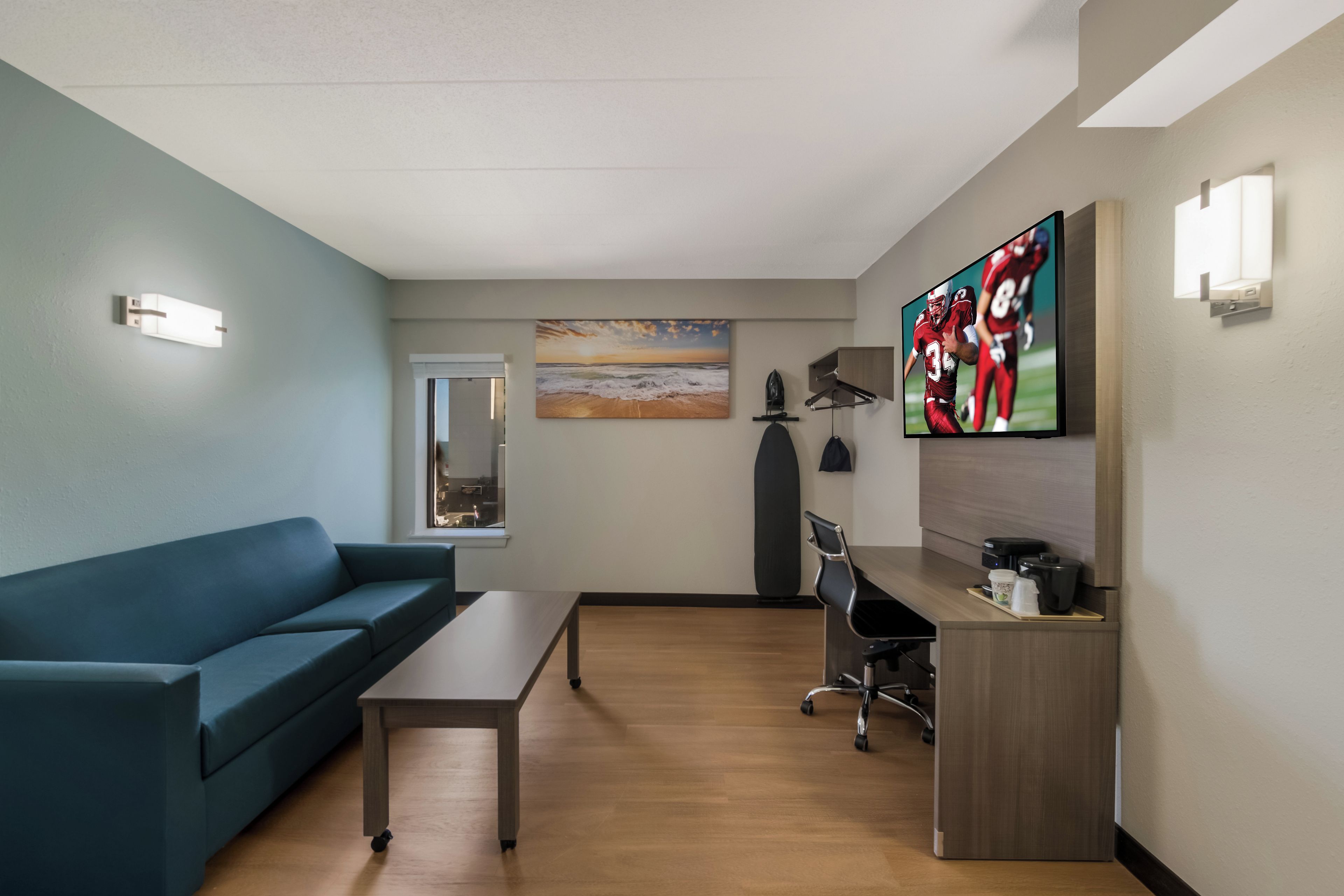 A modern hotel room with a blue sofa, a wooden coffee table, a desk with a computer, and a flat-screen TV mounted on the wall.