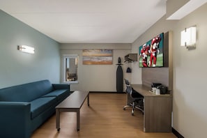 A modern hotel room with a blue sofa, a wooden coffee table, a desk with a computer, and a flat-screen TV mounted on the wall. - Red Roof Inn PLUS+ & Suites Virginia Beach – Seaside (Virginia Beach)